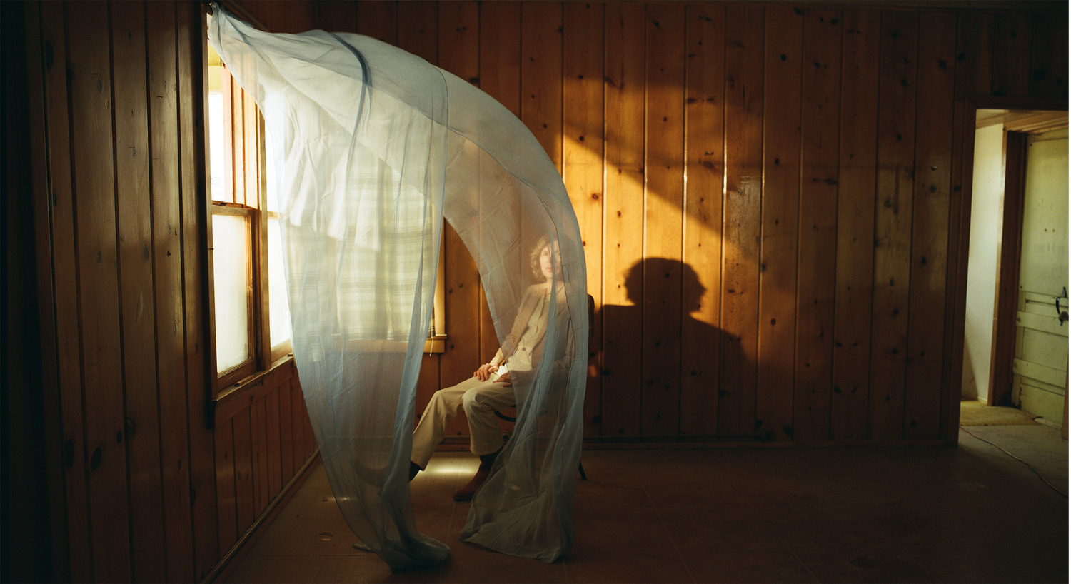 Beck sits inside a rustic room with wood-paneled walls, lit by warm sunlight, casting a long shadow on the wall and a white curtain blowing over Beck.
